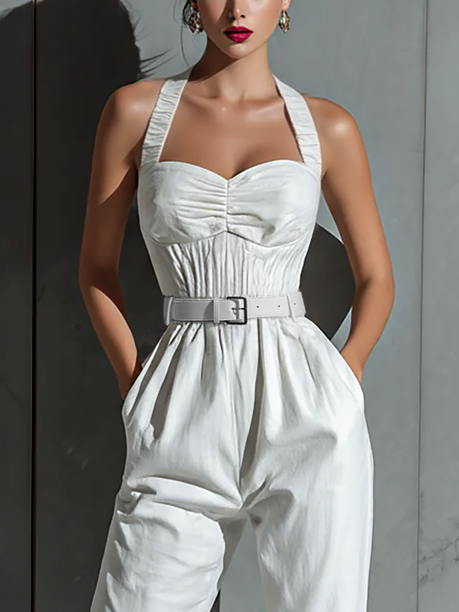 Ivory - A woman wearing a white textured linen halter-neck jumpsuit with a ruched bodice, a matching belted waist, and tapered trousers, standing against a grey marble wall.