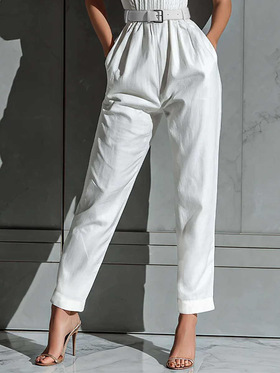 Ivory - A woman wearing a white textured linen halter-neck jumpsuit with a ruched bodice, a matching belted waist, and tapered trousers, standing against a grey marble wall.