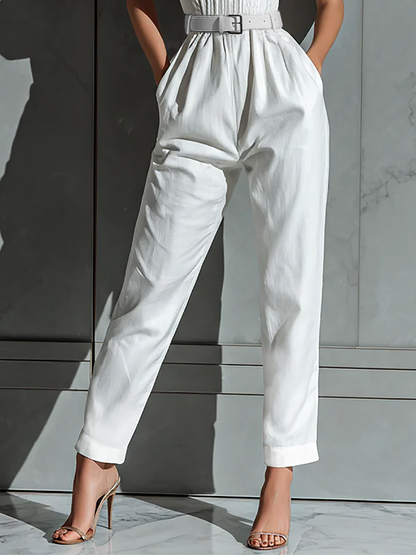 Ivory - A woman wearing a white textured linen halter-neck jumpsuit with a ruched bodice, a matching belted waist, and tapered trousers, standing against a grey marble wall.