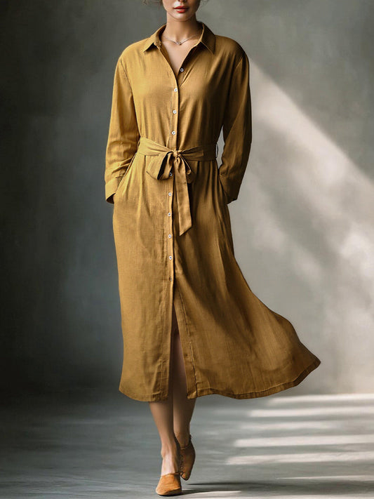 Mustard - A woman wearing a mustard-coloured, long-sleeve midi shirt dress made of a textured fabric. The dress features a pointed collar, button-down front, and a matching fabric tie belt at the waist.