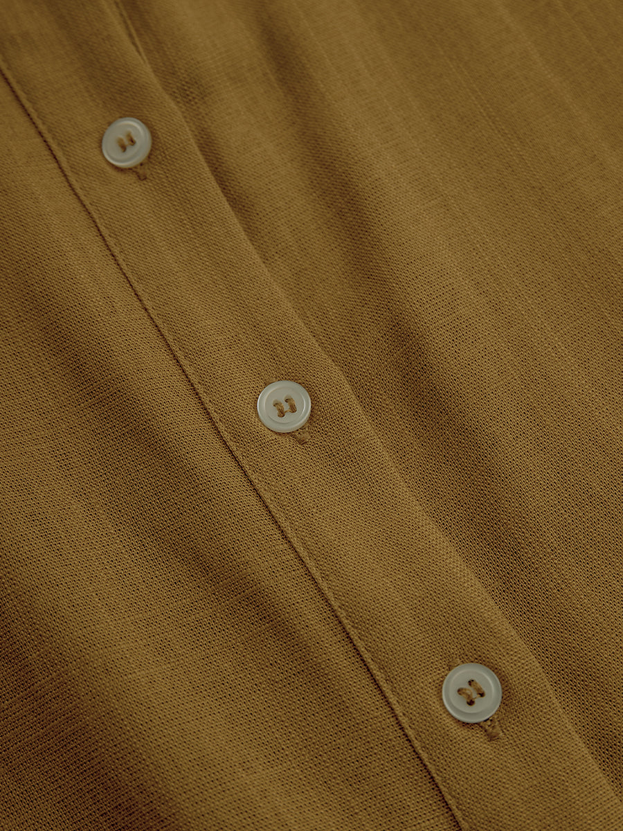 Mustard - A woman wearing a mustard-coloured, long-sleeve midi shirt dress made of a textured fabric. The dress features a pointed collar, button-down front, and a matching fabric tie belt at the waist.