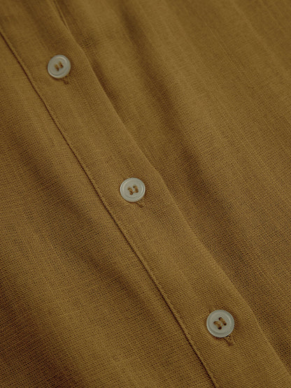 Mustard - A woman wearing a mustard-coloured, long-sleeve midi shirt dress made of a textured fabric. The dress features a pointed collar, button-down front, and a matching fabric tie belt at the waist.