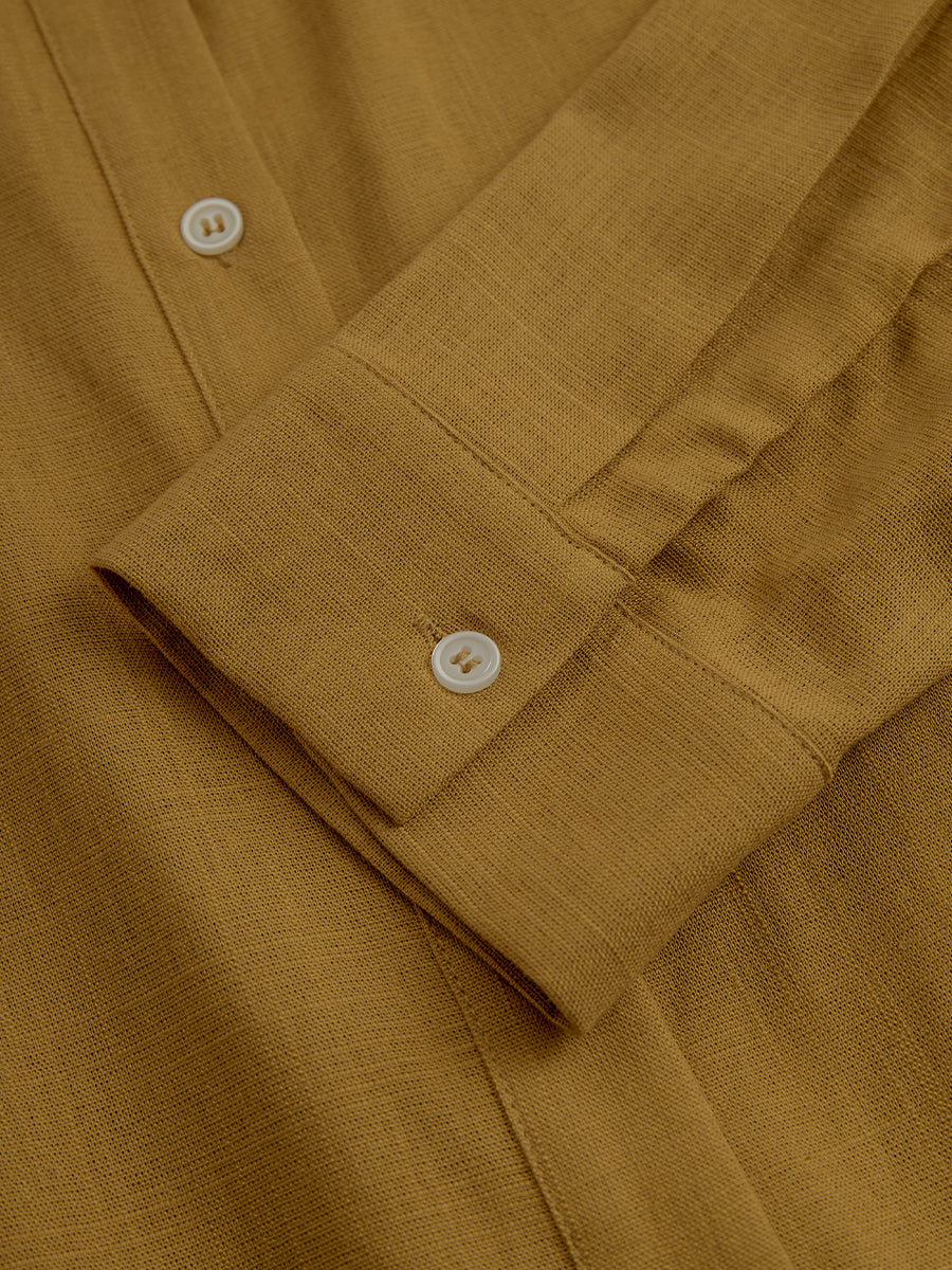 Mustard - A woman wearing a mustard-coloured, long-sleeve midi shirt dress made of a textured fabric. The dress features a pointed collar, button-down front, and a matching fabric tie belt at the waist.