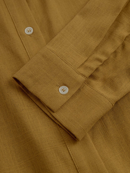 Mustard - A woman wearing a mustard-coloured, long-sleeve midi shirt dress made of a textured fabric. The dress features a pointed collar, button-down front, and a matching fabric tie belt at the waist.