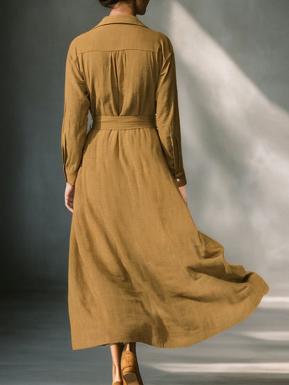 Mustard - A woman wearing a mustard-coloured, long-sleeve midi shirt dress made of a textured fabric. The dress features a pointed collar, button-down front, and a matching fabric tie belt at the waist.