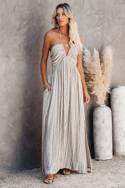 Grey - A woman posing in a neutral-toned, fine-striped halter maxi dress featuring a ruched bodice, deep V-neckline, and side splits, set against a minimalist concrete wall.