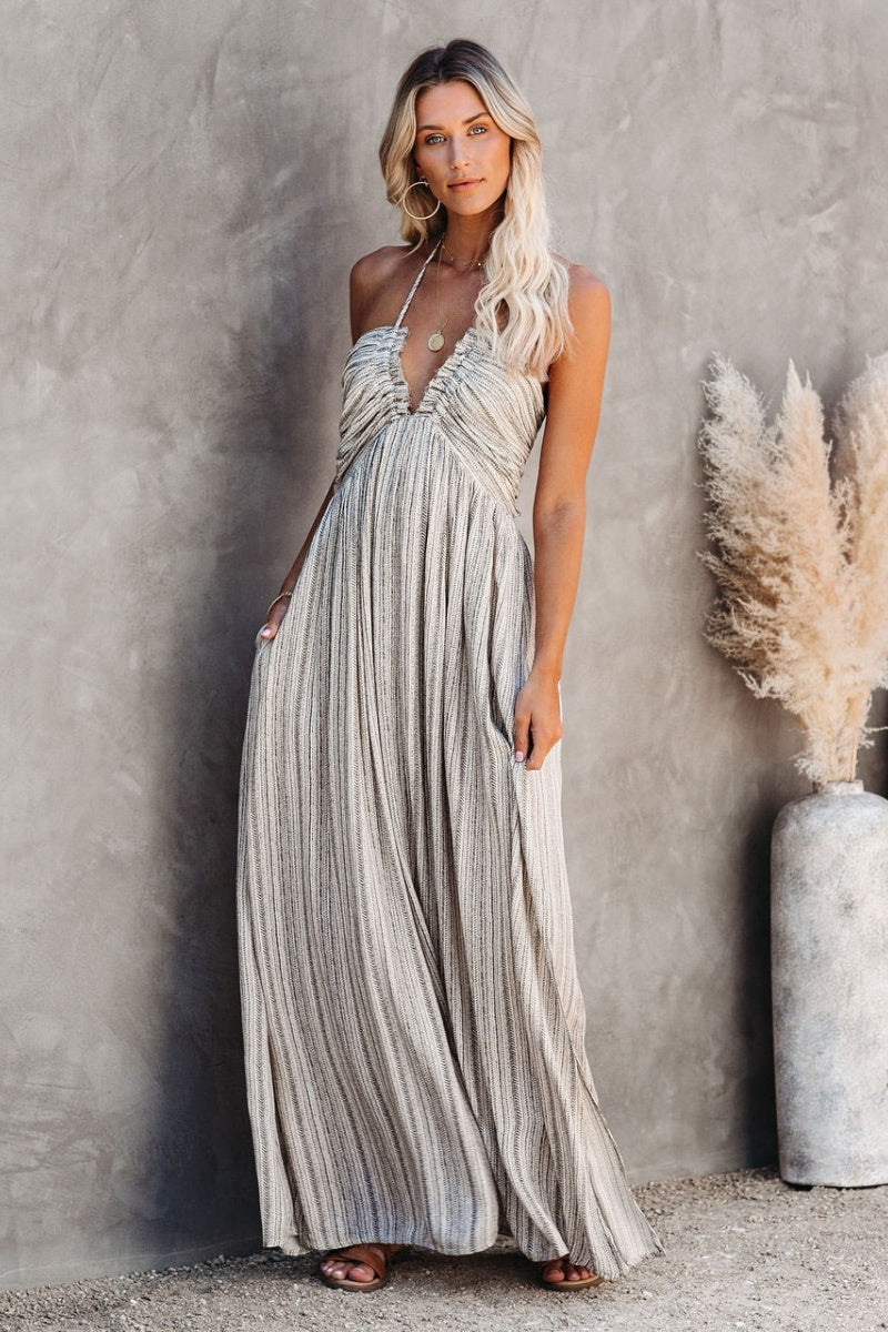 Grey - A woman posing in a neutral-toned, fine-striped halter maxi dress featuring a ruched bodice, deep V-neckline, and side splits, set against a minimalist concrete wall.