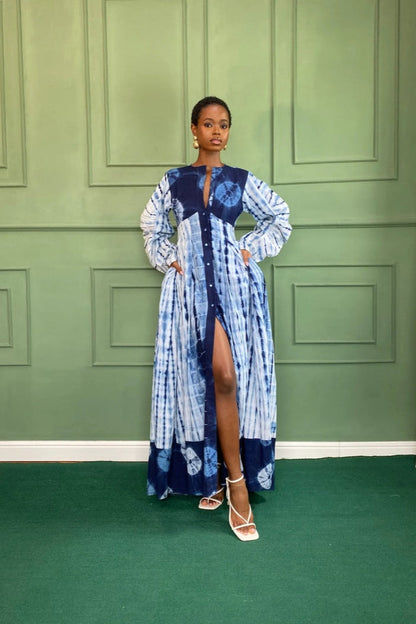 Blue - A woman wearing a long, flowing blue and white tie-dye maxi dress featuring a button-front closure, deep V-neckline, dramatic balloon sleeves, and side pockets, standing against a green paneled wall.