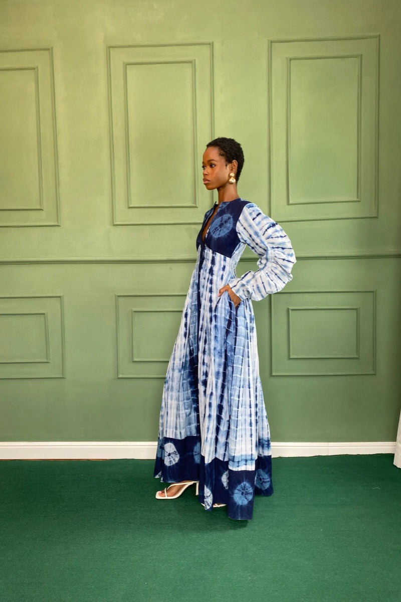 Blue - A woman wearing a long, flowing blue and white tie-dye maxi dress featuring a button-front closure, deep V-neckline, dramatic balloon sleeves, and side pockets, standing against a green paneled wall.