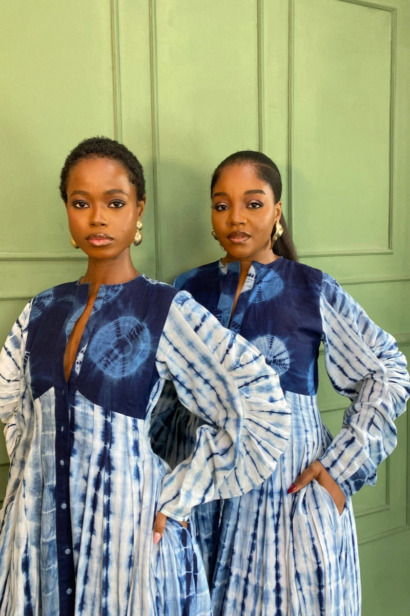Blue - A woman wearing a long, flowing blue and white tie-dye maxi dress featuring a button-front closure, deep V-neckline, dramatic balloon sleeves, and side pockets, standing against a green paneled wall.
