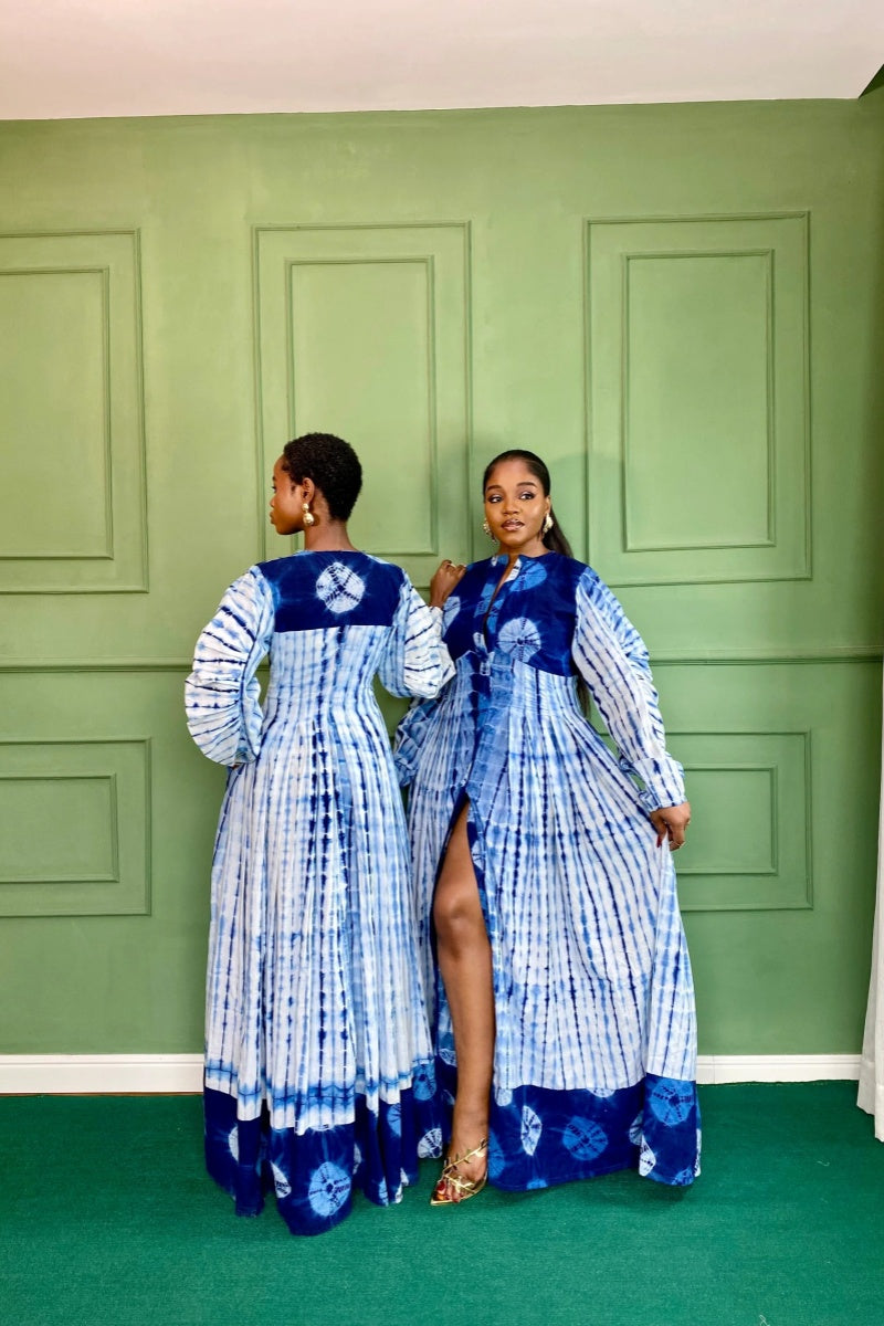 Blue - A woman wearing a long, flowing blue and white tie-dye maxi dress featuring a button-front closure, deep V-neckline, dramatic balloon sleeves, and side pockets, standing against a green paneled wall.
