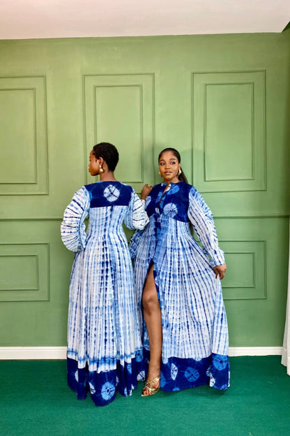 Blue - A woman wearing a long, flowing blue and white tie-dye maxi dress featuring a button-front closure, deep V-neckline, dramatic balloon sleeves, and side pockets, standing against a green paneled wall.