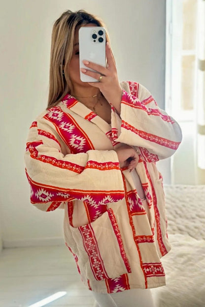 A woman wearing a relaxed, open-front kimono jacket featuring a vibrant pink and orange geometric Aztec print over a cream base, styled over a crochet crop top and shorts.